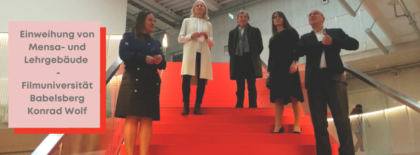 Zu sehen sind neben weiteren Personen, die Ministerin Manja Schüle und die Präsidentin der Filmuniversität, Susanne Stürmer auf einer roten Treppe im Neubau