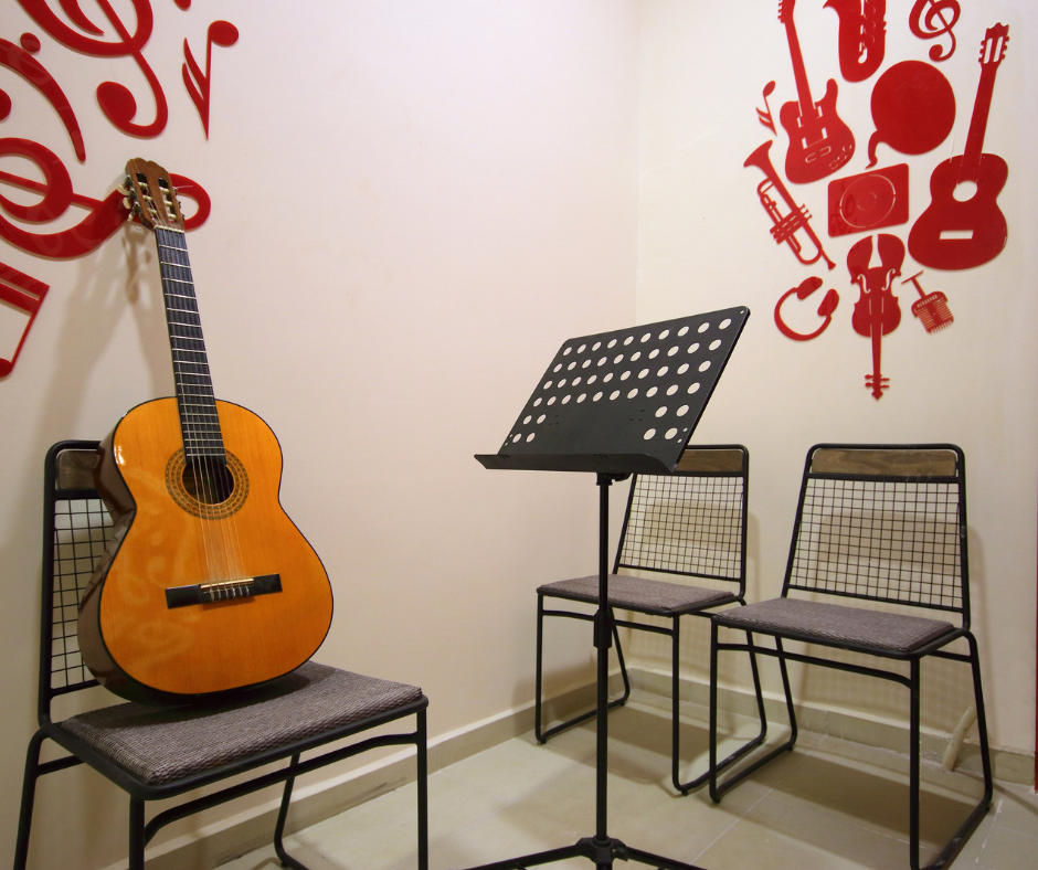 Zu sehen sind 3 Stühle, auf einem Stuhl steht eine Gitarre, im Hintergrund sind an der Wand Noten gezeichnet